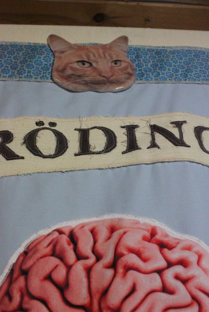A detail of a fabric stitched banner, with fragments of lettering of the word 'Schrödinger’s', showing a part of the printed fabric brain, lettering and fused glass 'cat deity'